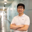 Man in white polo and glasses standing in a long indoor hallway, arms crossed