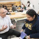 Male instructor speaking to student knitting purple yarn in a busy classroom workshop