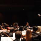 Orchestra on dark stage with conductor raising baton amid violinists under warm lights