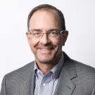 Smiling man with glasses, wearing a gray blazer and blue shirt, against a plain background.