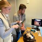 Two people work at a table with a monitor displaying a surgical procedure.