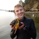 White male scientist in wet suit holds sea lemon and smiles at camera with ocean bay in background