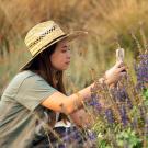 Woman sits in front of plants with purple flowers, taking a photo with her cell phone.