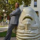 Chancellor Gary S. May, in suit, leans on "Eye on Mrak" Egghead sculpture.