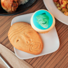 A small plate on a dining commons table holds a sugar cookie in the shape of an Egghead sculpture, Yin & Yang, and a smaller cookie with an edible image of another Egghead sculpture (Eye on Mrak, Fatal Laff)printed on it.