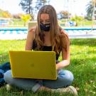 A student works on her laptop in a mask on the grass in front of a pool at UC Davis.