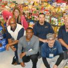 Seven employees pose in front of tables stacked with food donations.