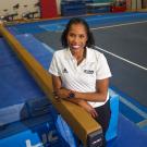 Female in white Aggie Athletics polo shirt, one arm atop balance beam in gym