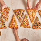 Hands reaching for slices of colorful pizza on a table with two pizza trays.