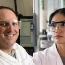 Two scientists in lab wearing safety goggles and white coats; man smiling, woman serious.