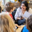 Teacher seated, with dark hair and white blouse, talks to kindergartners 