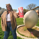 Aggie student stands in front of an Egghead sculpture while holding an Egghead Collectible Cup available at Spokes for a limited time