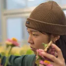 Person in brown beanie tending pink and yellow flowers by a window, photograph