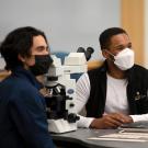 Two students wearing face coverings sit at table in laboratory.