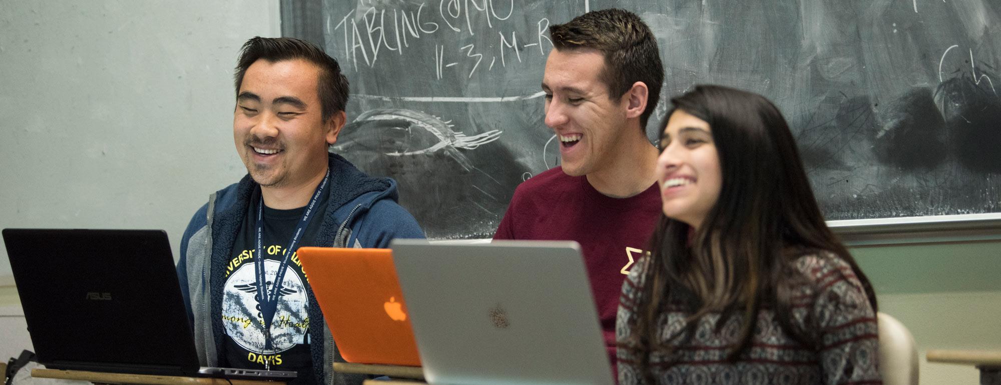 three students sitting in front of a chalkboard with laptops laughing in a classroom