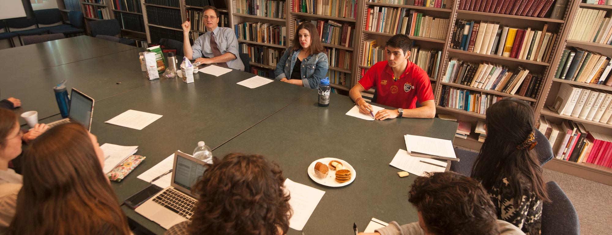 students and a professor sitting around a large table in a room with many books discussing an assignment