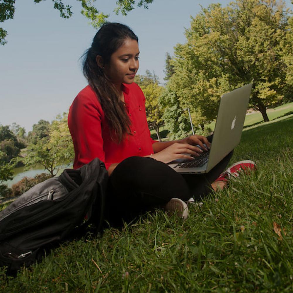 A female student in red reads from her laptop on the UC Davis quad