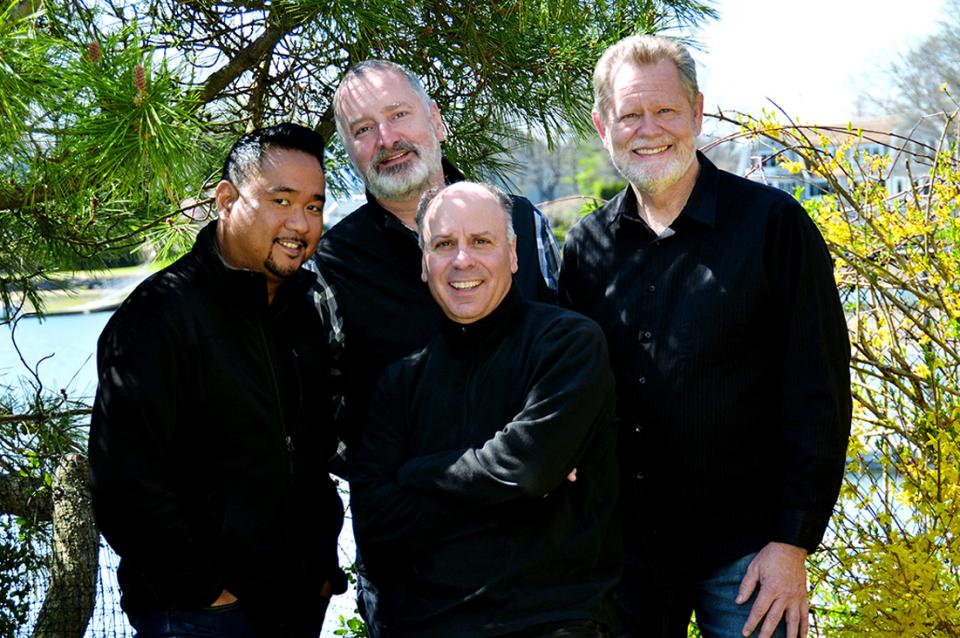 Members of the quartet wearing mosly black, staning outside near a tree and other greenery.