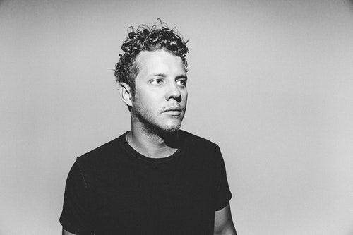 Anderson East wearing a black t-shirt, standing in front of a gray background.
