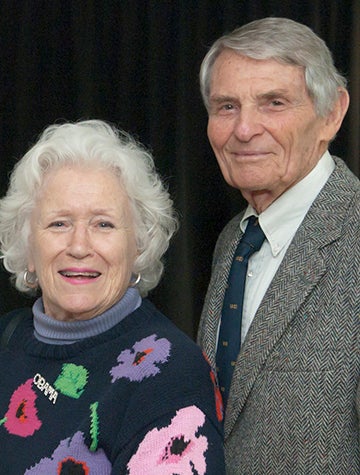 Woman with white hair in flowered sweater stands next to main with gray hair in suit and tie
