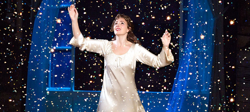 A young woman cast member smiling onstage with her hands raised, surrounded by fairy dust.