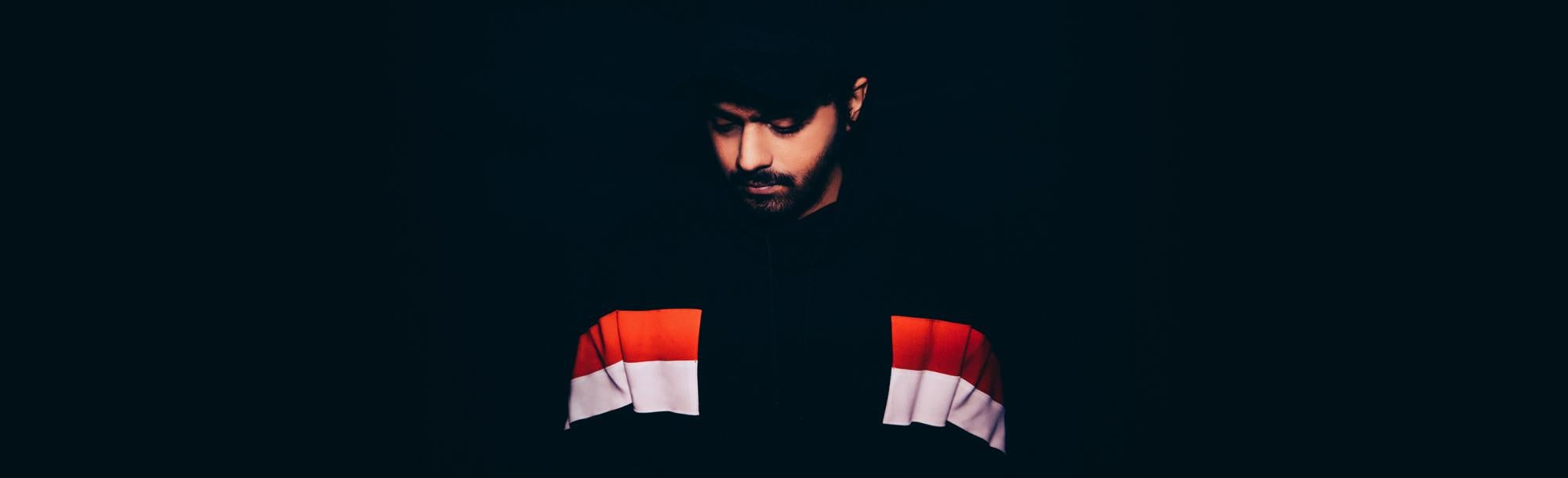 Jai Wolf looking downward, wearing mostly black clothing and standing against a black background.