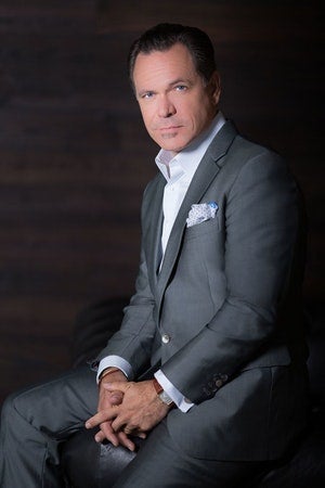 Kurt Elling sitting with his hands crossed, wearing a gray suit and white dress shirt, staring into the camera.
