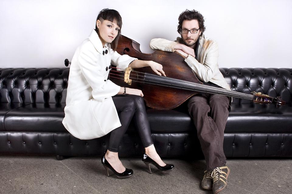 The two members of the group sitting on a black leather couch with a double-bass.