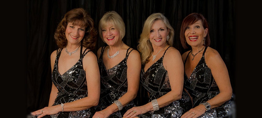 The members of the New Chordettes wearing matching dresses and posing in front of a dark background.